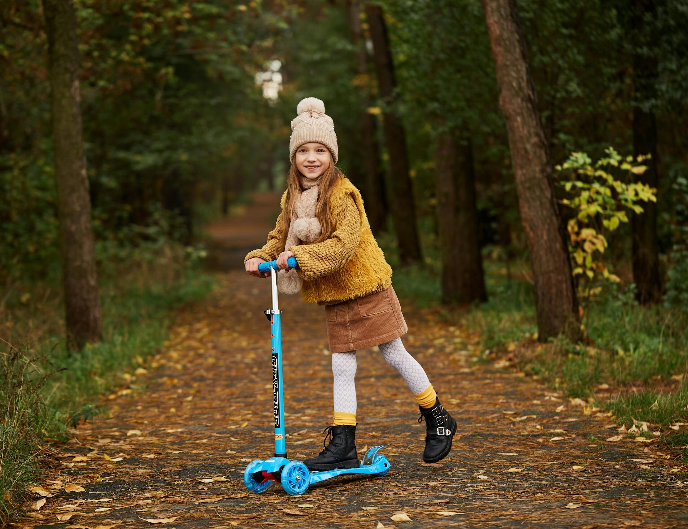 Happy child in winter clothes riding a scooter along a forest path in autumn.